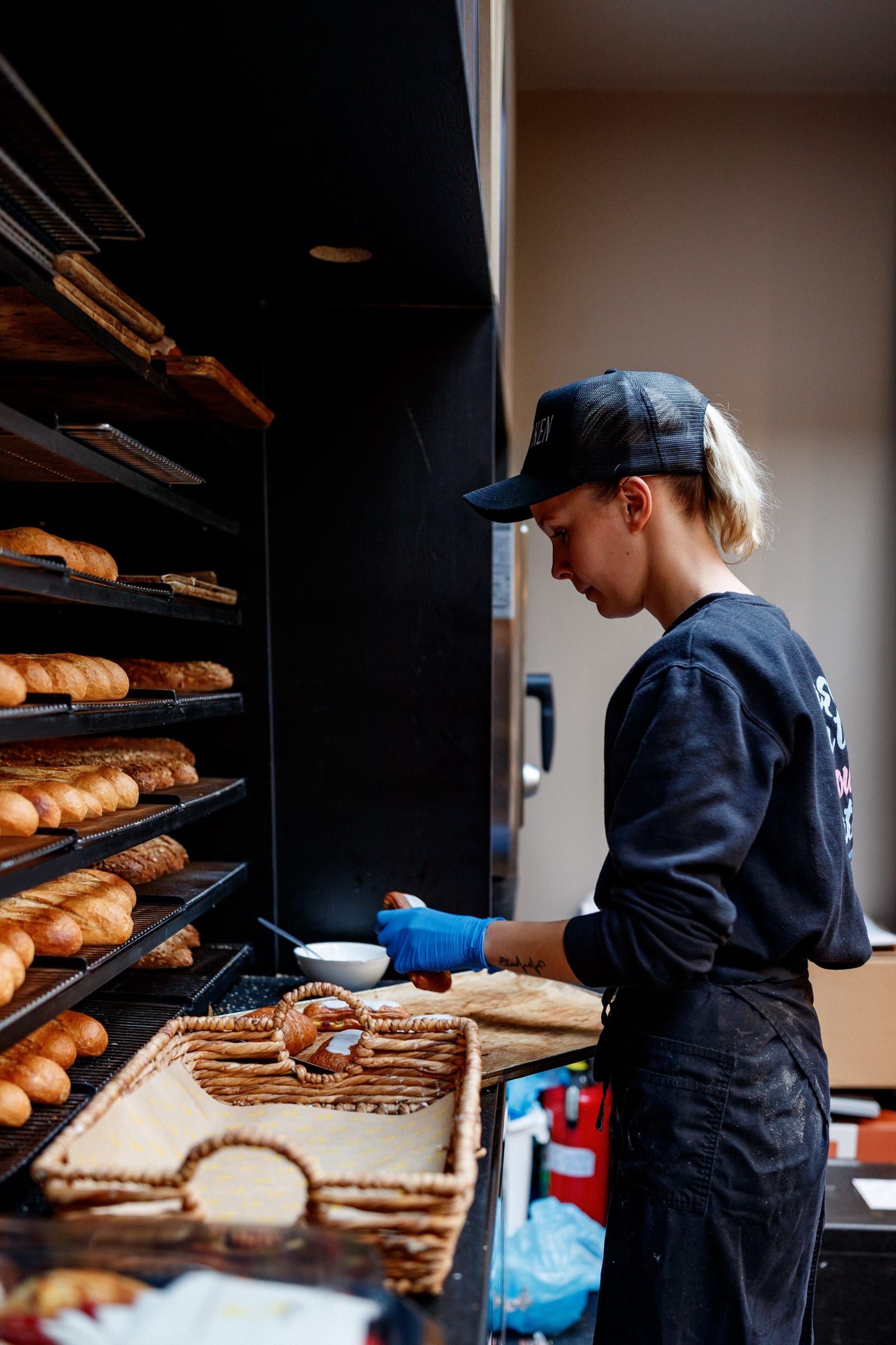 Medewerker met zwarte pet maakt broodjes klaar naast rekken met baguettes