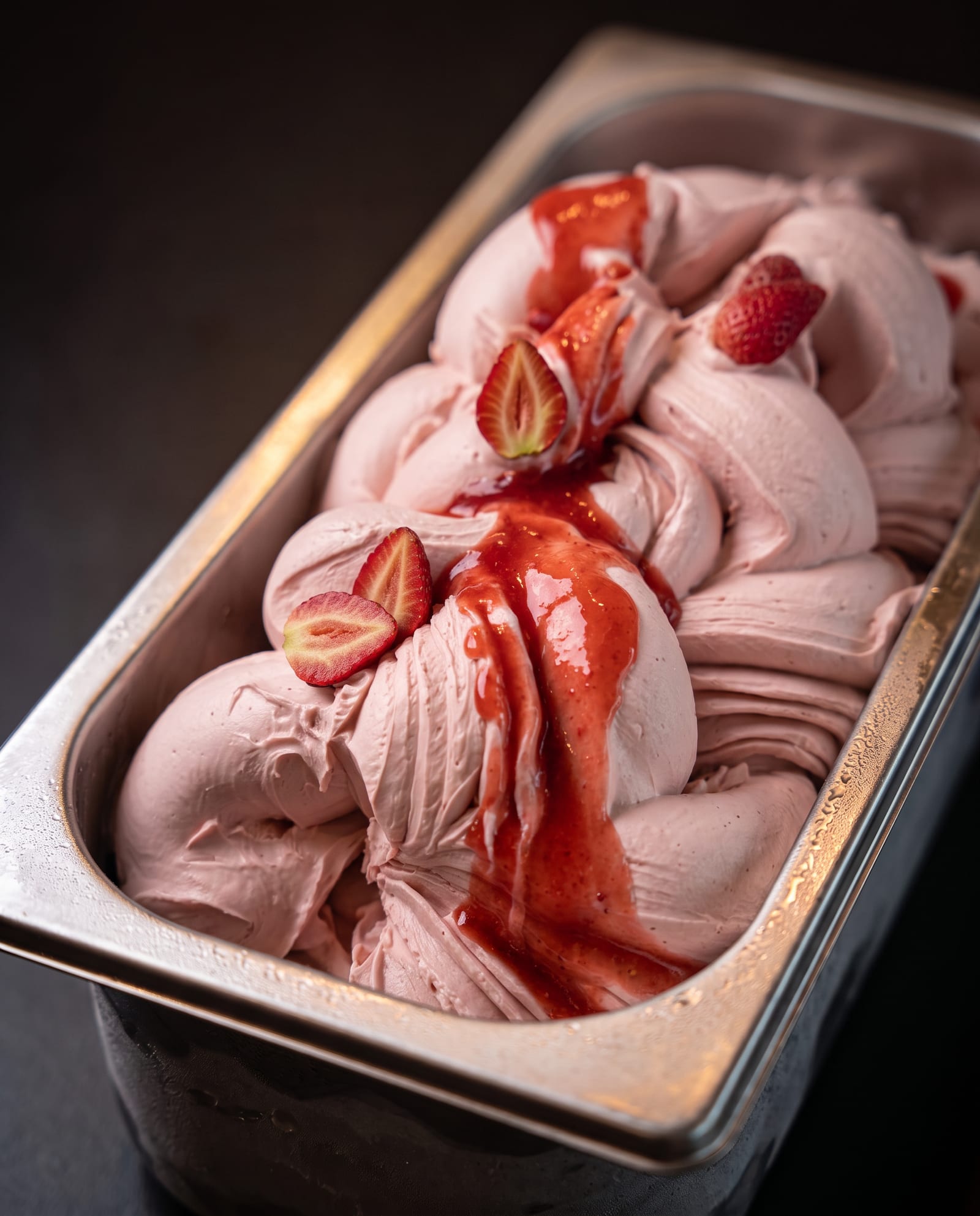 Stalen ijsbak schuin van boven op een donkere achtergrond, gevuld met roze aardbeienijs met halve verse aardbeien en aardbeiensaus