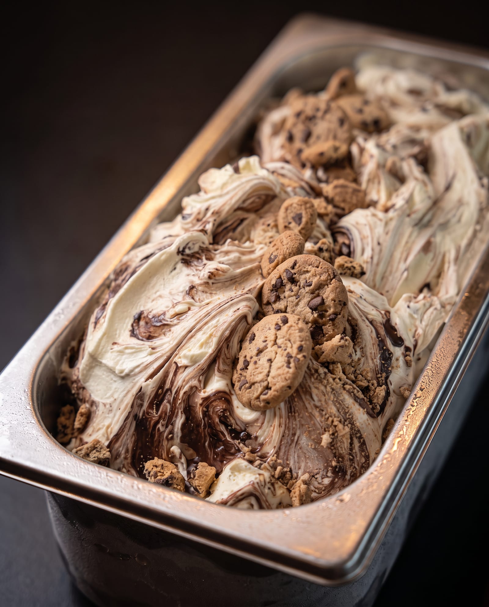 Stalen ijsbak schuin van boven op een donkere achtergrond, gevuld met crèmekleurig ijs met een chocoladeswirl en chocolate-chip-cookies