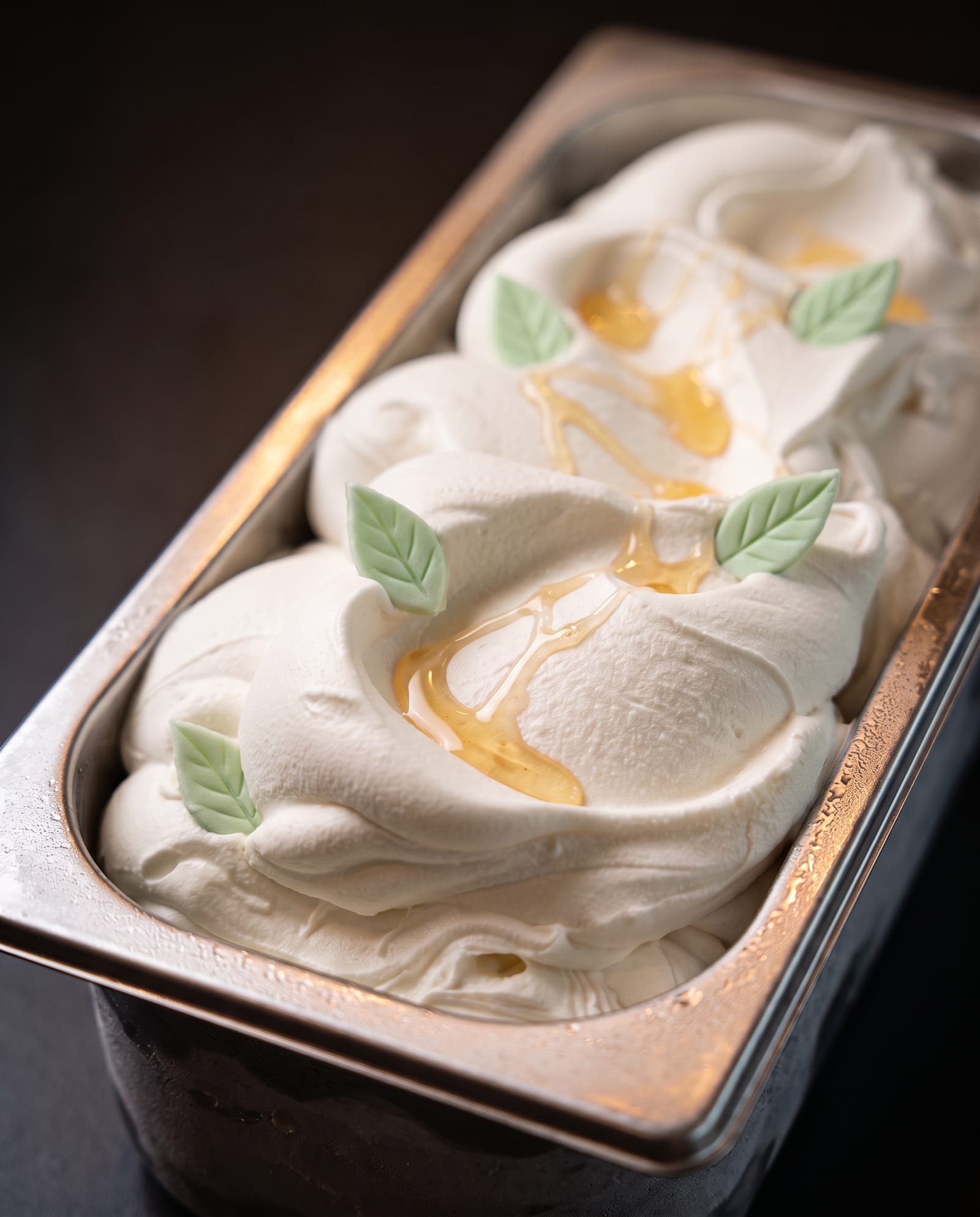 Stalen ijsbak schuin van boven op een donkere achtergrond, gevuld met crèmekleurig caramelijs met slierten caramelsaus en lichtgroene suikerblaadjes als decoratie
