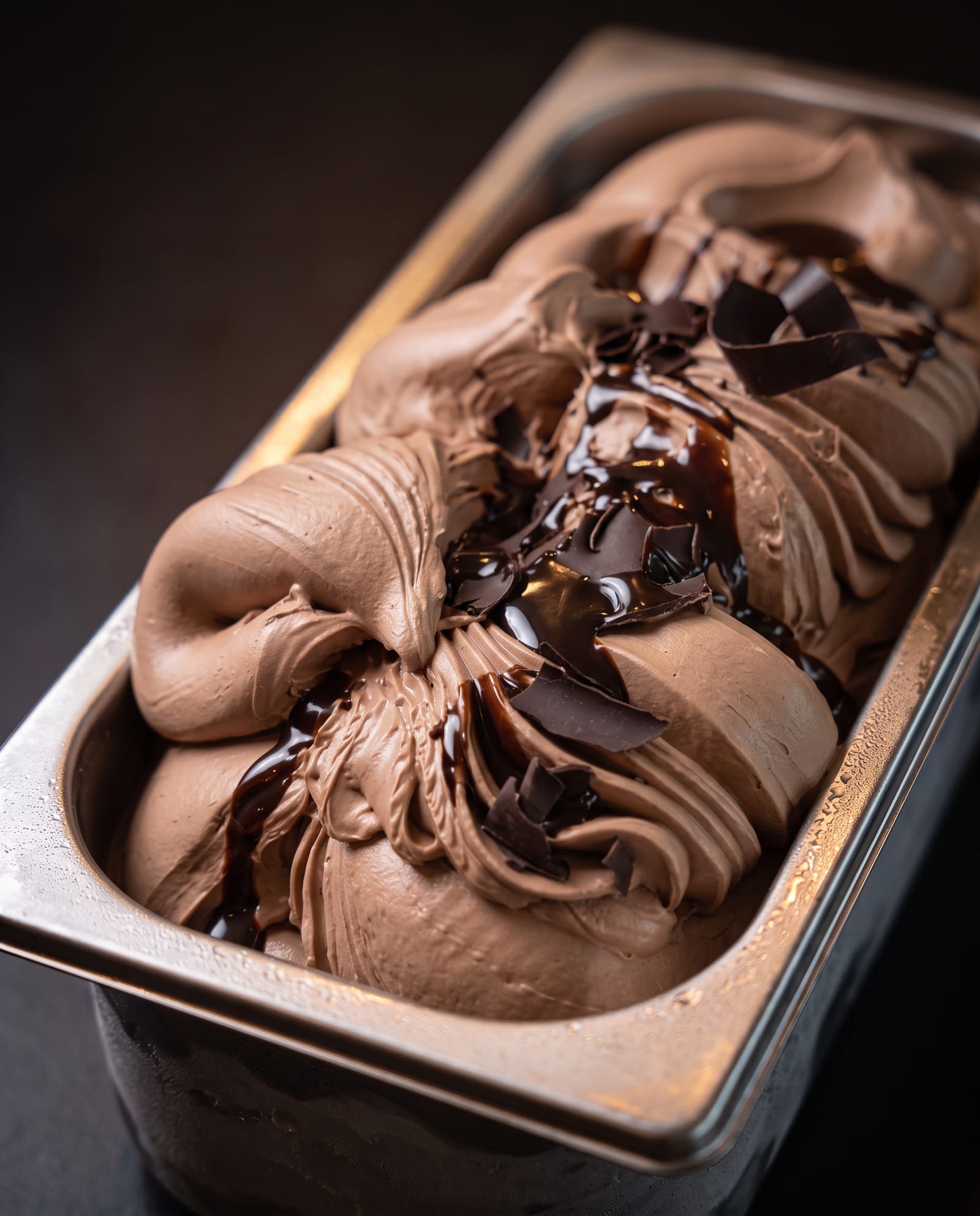 Stalen ijsbak schuin van boven op een donkere achtergrond, gevuld met melkchocolade-ijs met chocoladekrullen en glanzende chocoladesaus