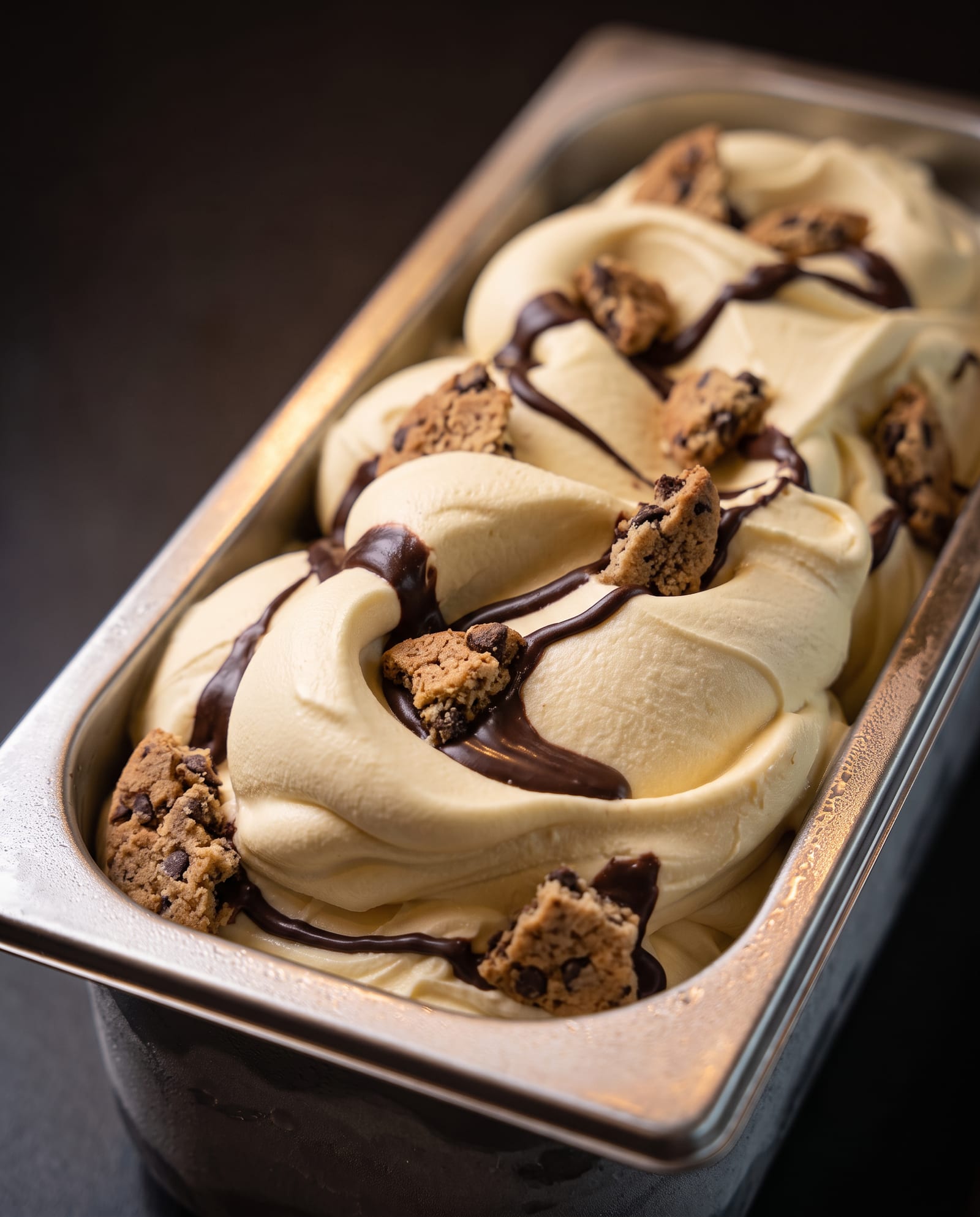 Stalen ijsbak schuin van boven op een donkere achtergrond, gevuld met geel ijs met een chocoladeswirl en stukjes American cookie