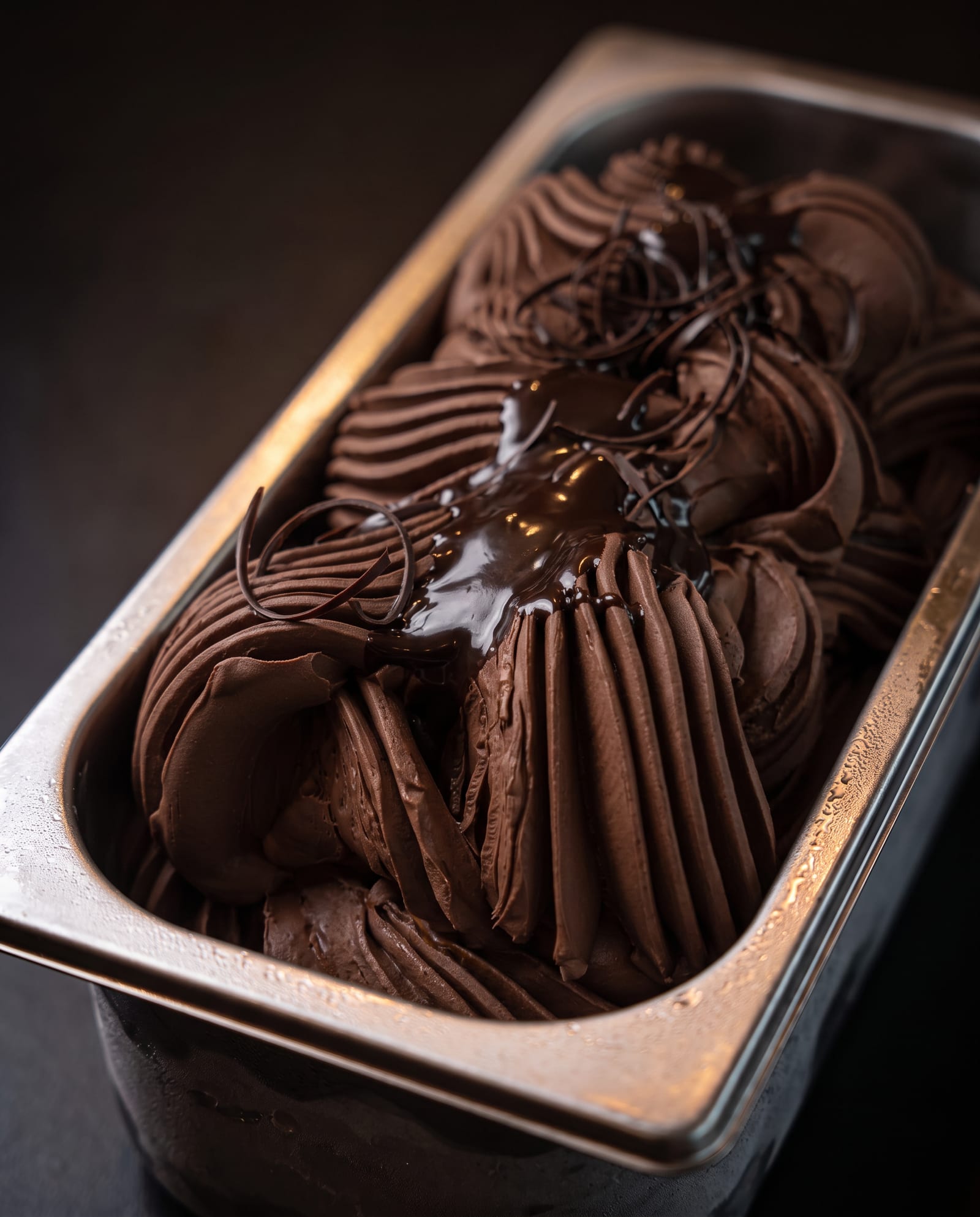 Stalen ijsbak schuin van boven op een donkere achtergrond, gevuld met donker fondant-chocolade-ijs in diepe ribbels met glanzende pure chocoladesaus