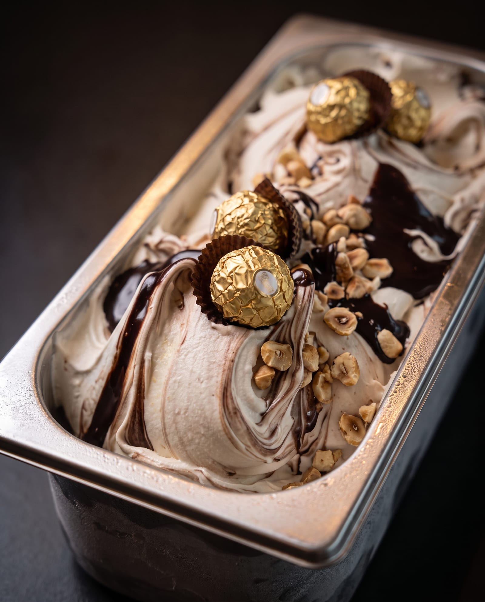 Stalen ijsbak schuin van boven op een donkere achtergrond, gevuld met licht hazelnootijs met een chocoladeswirl, hazelnoten en pralines in goudfolie