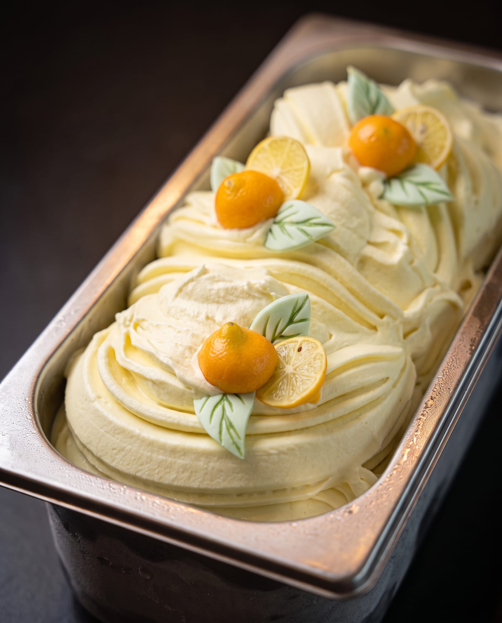 Stalen ijsbak schuin van boven op een donkere achtergrond, gevuld met lichtgele limoncellosorbet met citroenschijfjes, kleine oranje citrusvruchtjes en lichtgroene suikerblaadjes