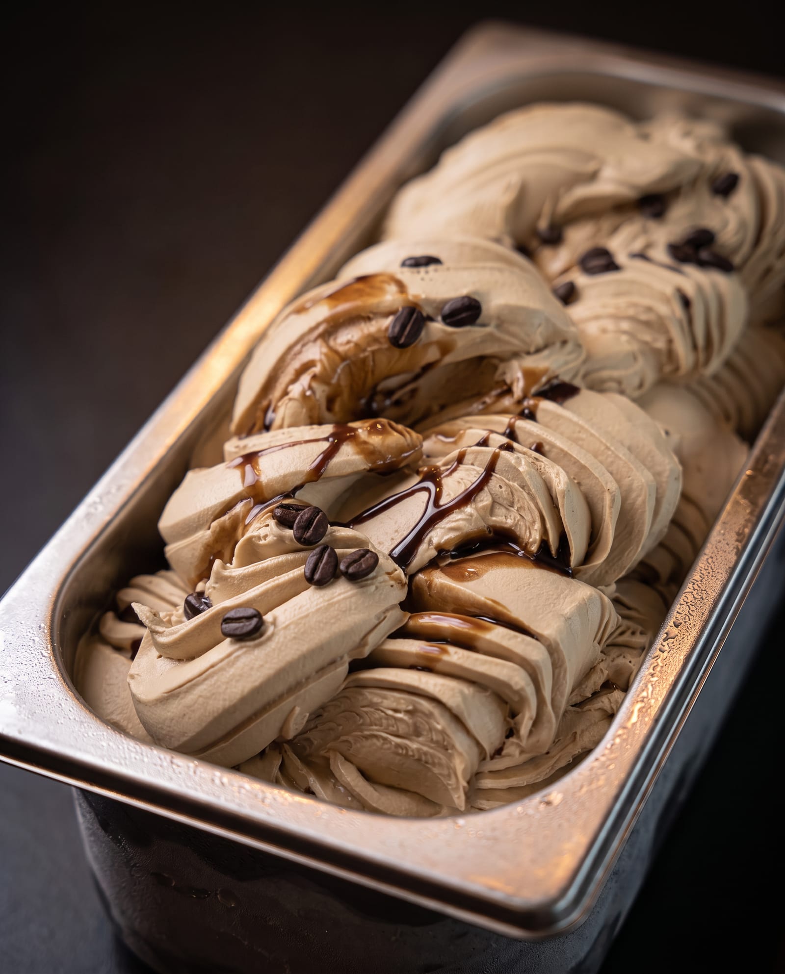 Stalen ijsbak schuin van boven op een donkere achtergrond, gevuld met lichtbruin koffie-ijs in swirls met koffiebonen en dunne strepen chocoladesaus