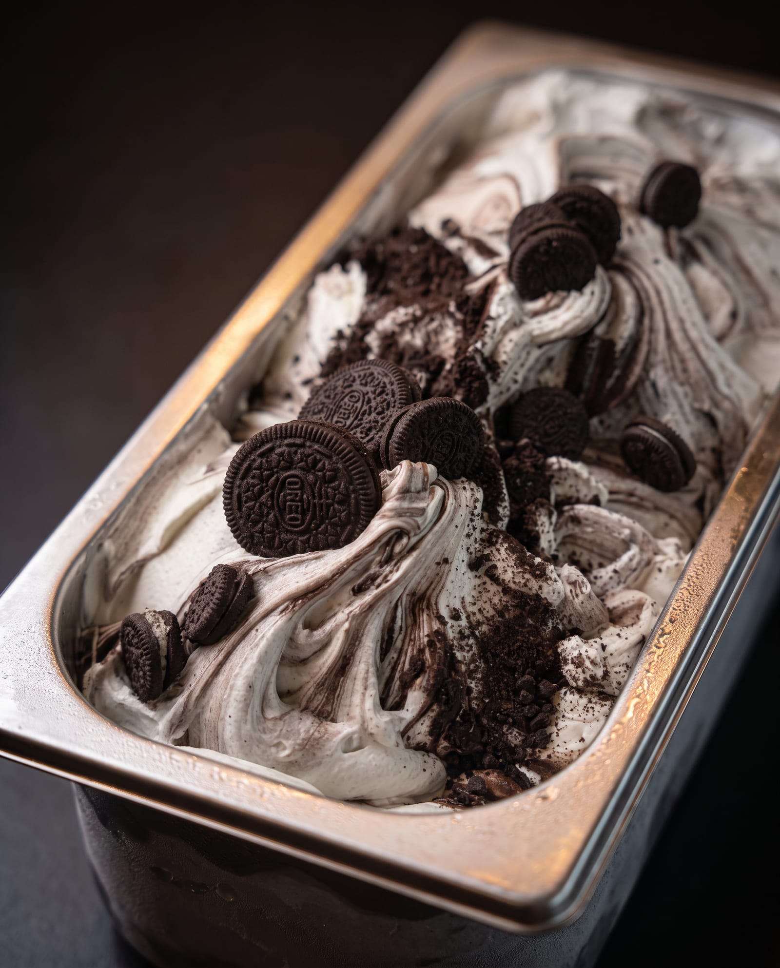 Stalen ijsbak schuin van boven op een donkere achtergrond, gevuld met wit-grijs ijs gemarmerd met koekkruimels, met hele en gebroken Oreo-koekjes