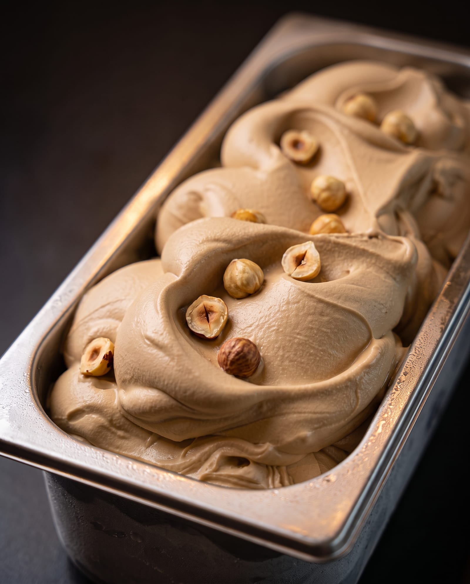 Stalen ijsbak schuin van boven op een donkere achtergrond, gevuld met koffie-caramelkleurig pralinéijs met hele en gehakte hazelnoten