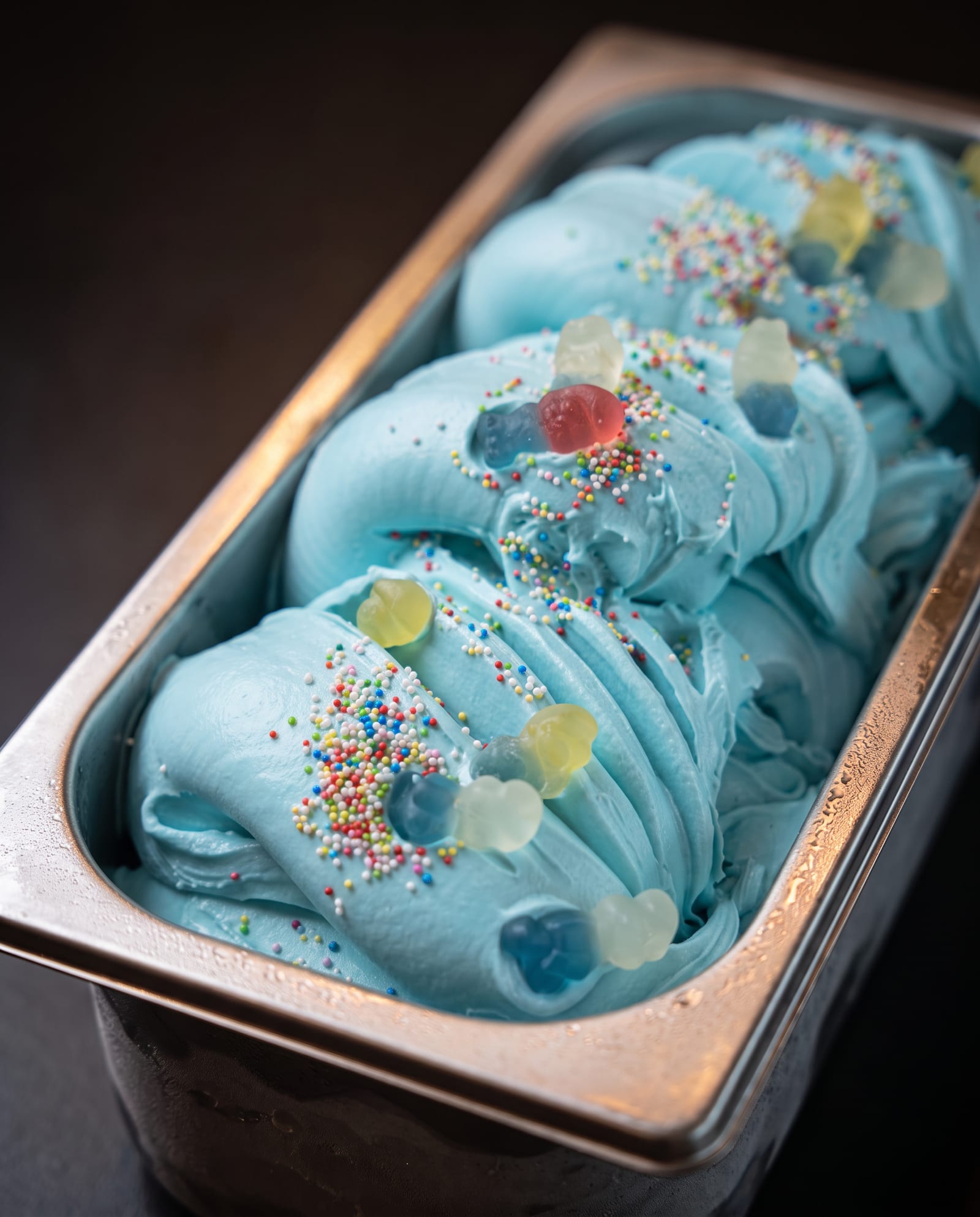 Stalen ijsbak schuin van boven op een donkere achtergrond, gevuld met hemelsblauw smurfenijs met gekleurde suikerstrooisels en gummibeertjes