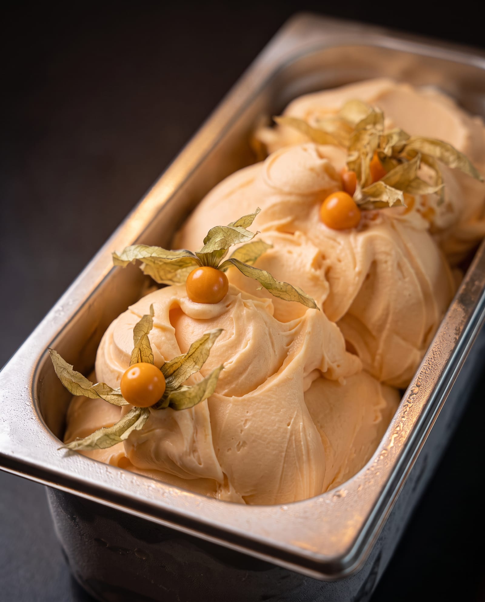 Stalen ijsbak schuin van boven op een donkere achtergrond, gevuld met abrikooskleurige tropical sorbet in swirls met physalis (goudbessen)