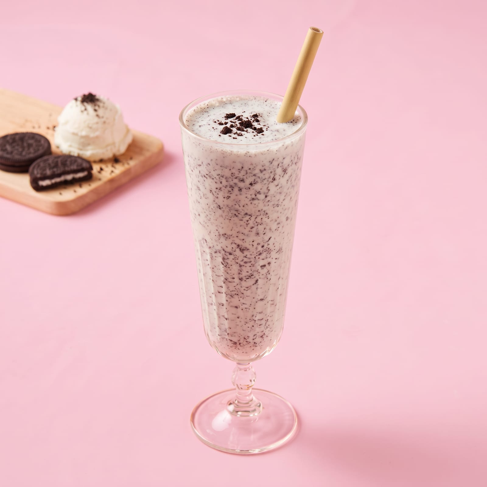 Milkshake Oreo met koekkruimels in een hoog glas op voet, met bamboe rietje, op een roze achtergrond; op een houten plankje een bol vanille-ijs en koekjes